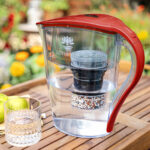 Eine rote Wasserfilter-Karaffe für die ganze Familie steht gefüllt mit gefiltertem Wasser auf einem Holztisch im Garten, daneben ein Glas mit frischem Wasser.