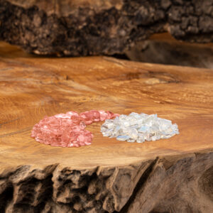 Zwei verschiedene Kristallstein-Mischungen, eine rötlich und eine weißlich-schimmernd, liegen auf einer rustikalen Holzoberfläche.
