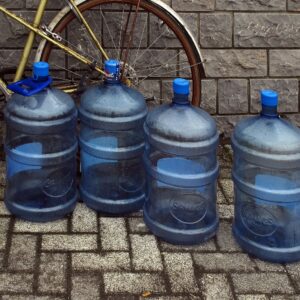 Vier große, blaue Wasserkanister stehen als Trinkwasservorrat aufgereiht auf einem gepflasterten Boden vor einer grauen Steinmauer.