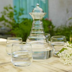 Eine formschöne Glaskaraffe und vier passende Trinkgläser, gefüllt mit reinem Wasser, stehen auf einem Holztisch in einem sommerlichen Garten.