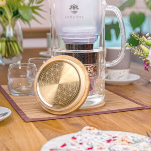 Eine Glas-Wasserfilterkanne mit einem goldenen Deckel, verziert mit der Blume des Lebens, steht auf einem liebevoll gedeckten Esstisch mit Blumen.
