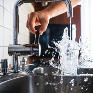 Frisches Wasser spritzt aus einem Glas, das unter einem laufenden Küchen-Wasserhahn überläuft.