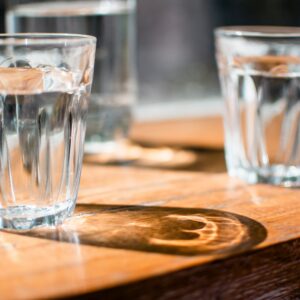Drei Gläser mit klarem Wasser auf einem Holztisch, das vorderste Glas im Fokus.