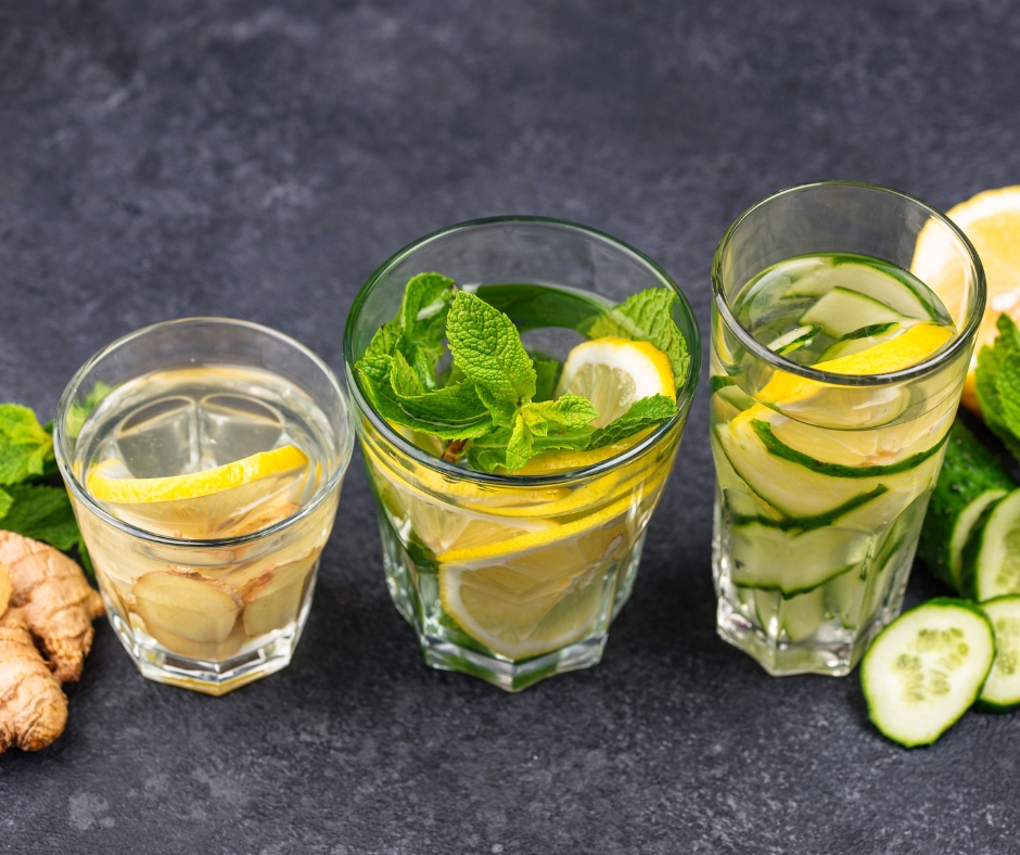 Drei Gläser mit gefiltertem Wasser, verfeinert mit frischen Zutaten wie Zitrone, Ingwer, Gurke und Minze auf einem dunklen Tisch.