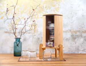 Ein Wasserfilter aus Glas und Bambus von Lotus Vita steht auf einem Holztisch, daneben zwei Gläser und eine Vase mit Hagebuttenzweigen.