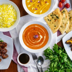 Auf einem gedeckten Tisch stehen gelber Reis, zwei Schalen Curry und Kräuterfladenbrot neben weiteren Speisen.