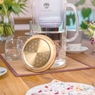 Eine Glas-Wasserfilterkanne mit einem goldenen Deckel, verziert mit der Blume des Lebens, steht auf einem liebevoll gedeckten Esstisch mit Blumen.
